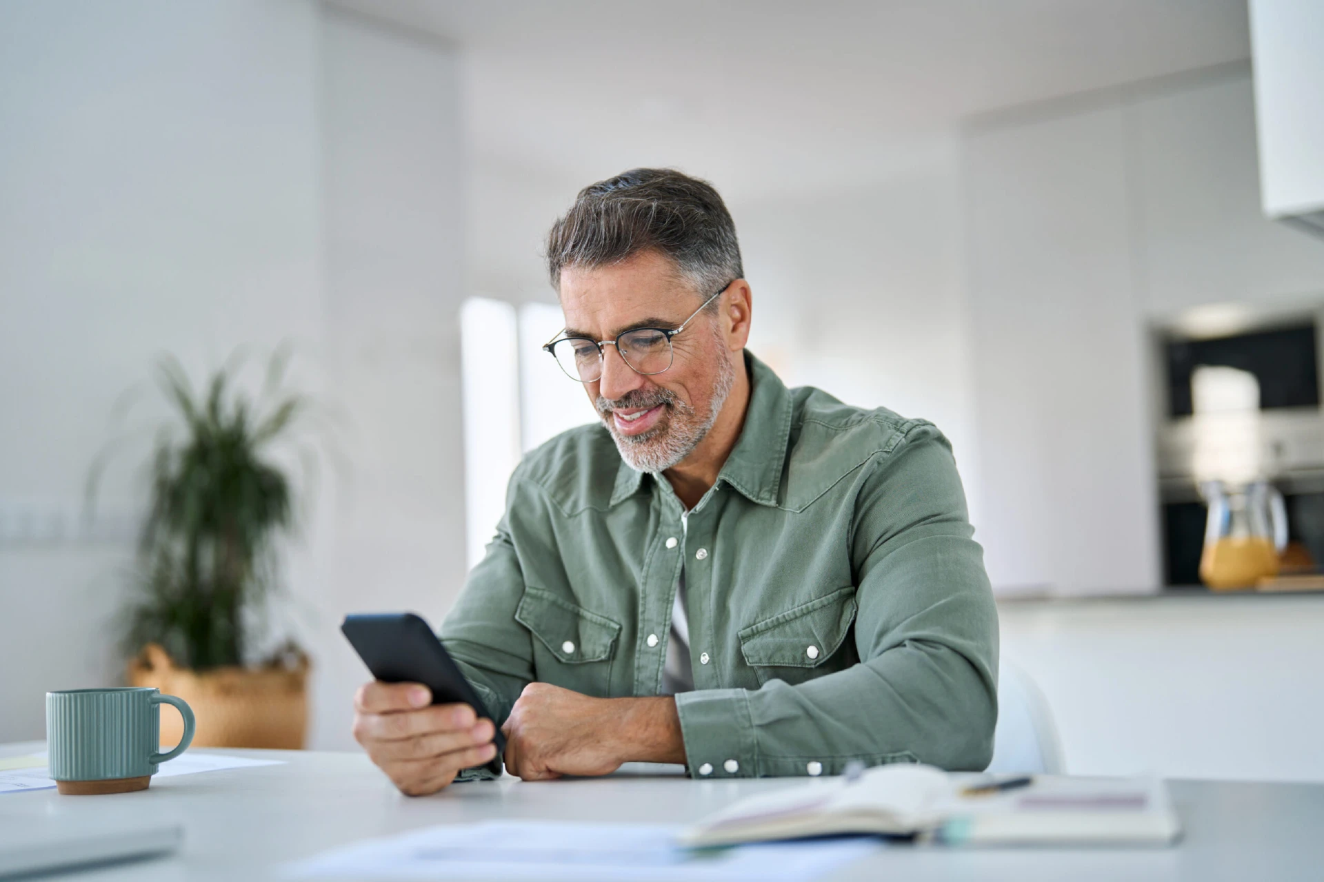 Happy mature senior man using mobile phone sitting at kitchen table. Smiling old middle aged customer holding smartphone scrolling buying online doing shopping,reading news in smartphone at home.